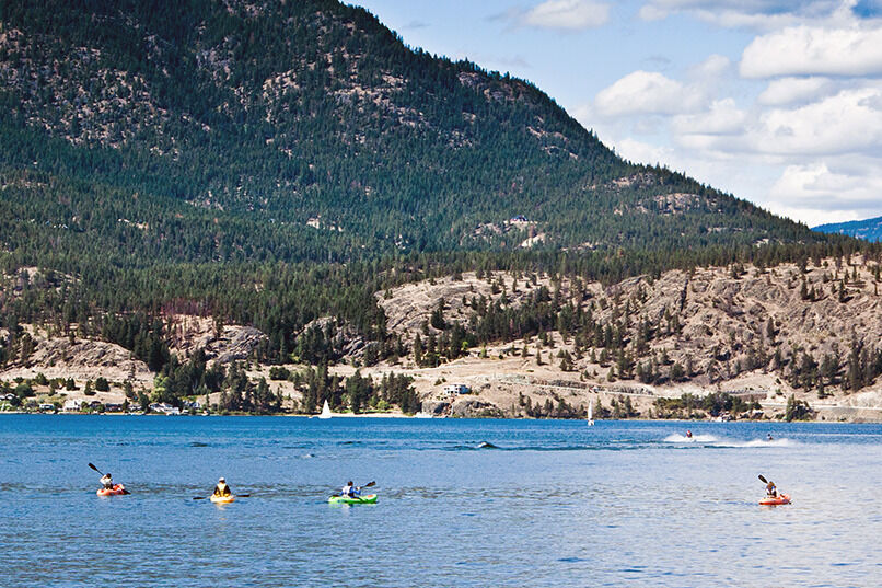 Four kayakers on Okanagan Lake with the rocky, pine-covered hills of West Kelowna rising behind them. This is a typical summer day on the water.